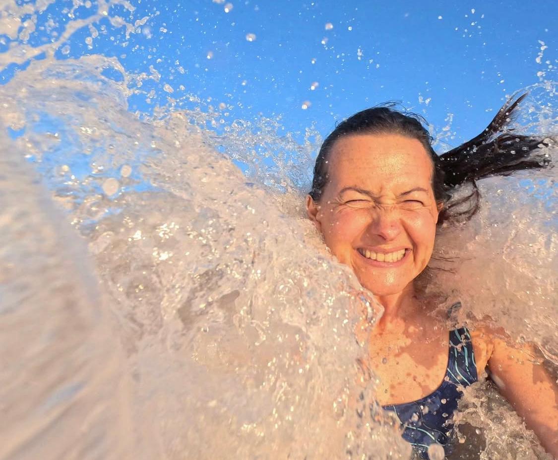 A selfie of El Etherington as a wave crashes over her, she is grinning from ear to ear as her hair blows wildly about - blue sky above and a sunset glow.